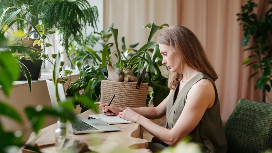 Woman taking notes at a laptop at a wooden desk surrounded by indoor plants